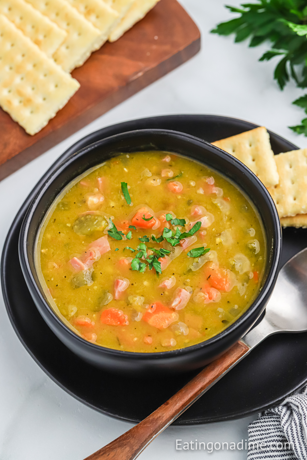 A black bowl of split pea soup on a plate with a spoon and saltine crackers