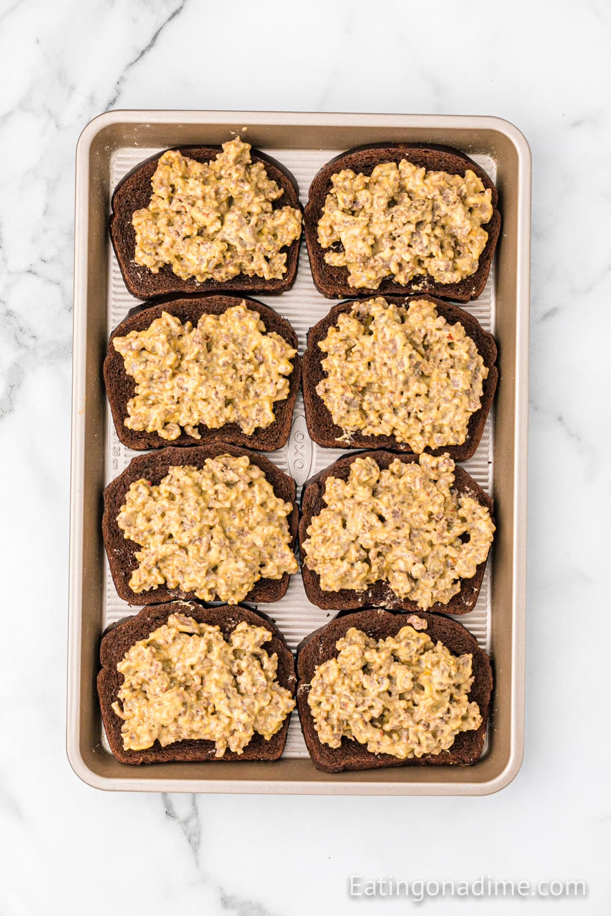 A baking sheet with eight slices of dark bread topped with a savory Hanky Panky Recipe mixture of ground meat and cheese, arranged in two rows on a white marble surface.