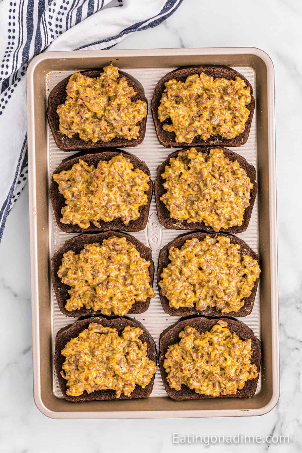 A baking sheet with eight slices of dark bread, each topped with a savory Hanky Panky Recipe ground meat and cheese mixture, is set on a marble surface next to a striped kitchen towel.