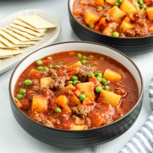 A bowl of hearty crock pot ground beef stew with chunks of potato, carrots, peas, and ground beef in a tomato-based broth, served next to a plate of saltine crackers.