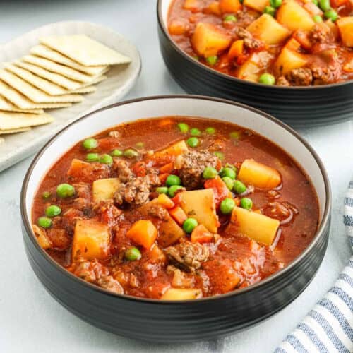 A bowl of hearty crock pot ground beef stew with chunks of potato, carrots, peas, and ground beef in a tomato-based broth, served next to a plate of saltine crackers.
