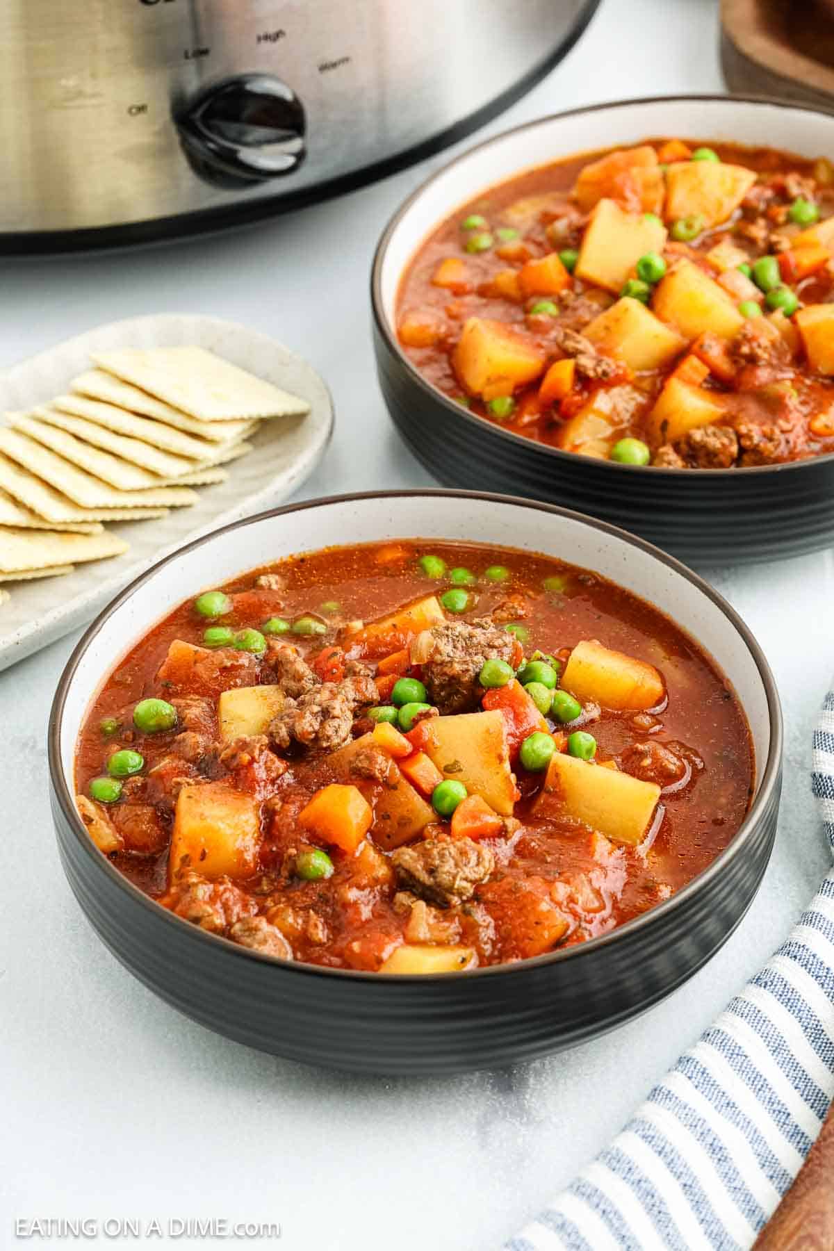 Two bowls of hearty crock pot ground beef stew filled with potatoes, carrots, peas, and beef in a rich tomato broth sit beside saltine crackers. A slow cooker is visible in the background, perfect for this easy stew recipe.