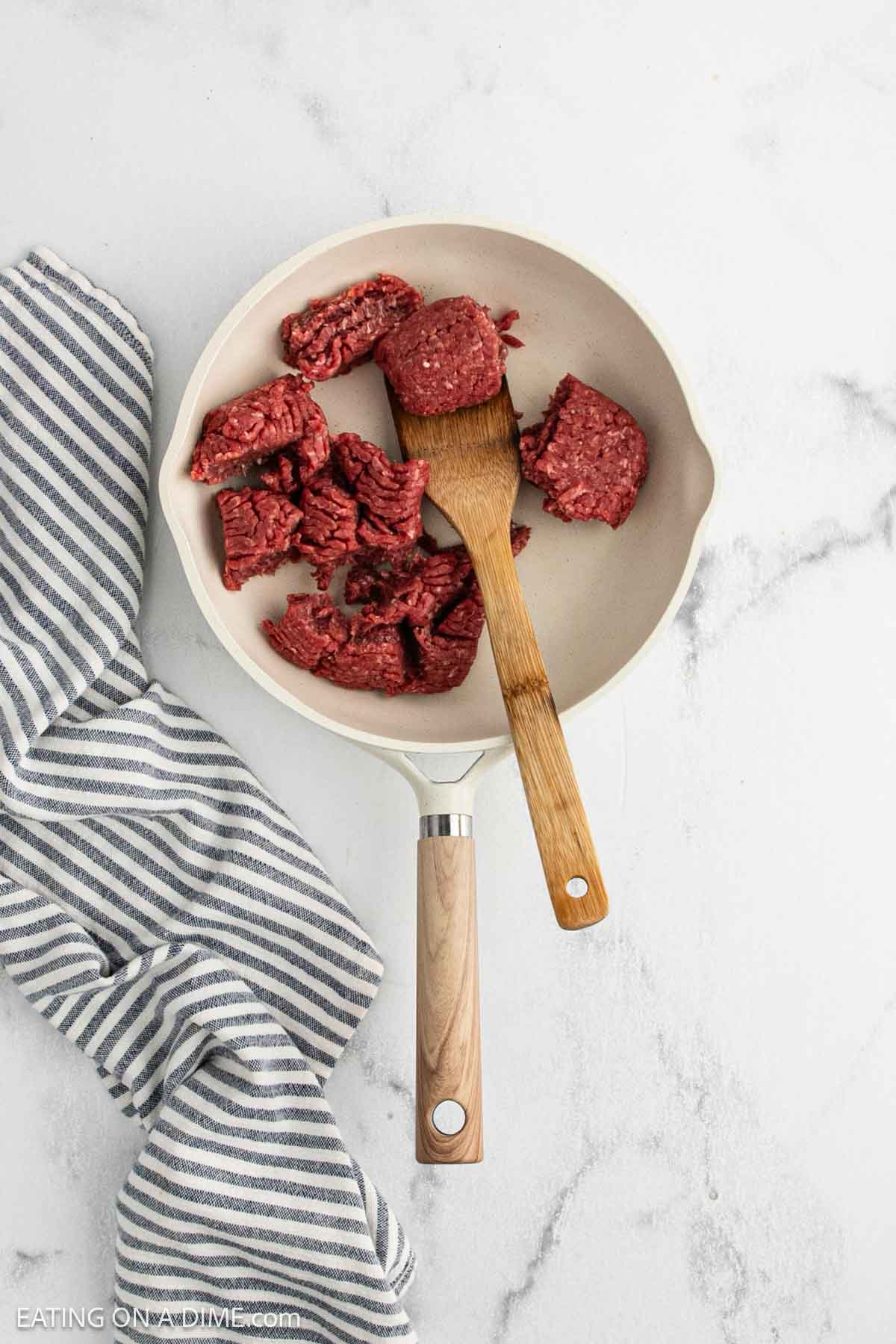 A white skillet with raw ground beef and a wooden spatula inside, placed on a white marble surface next to a folded blue and white striped kitchen towelโperfect for starting your crock pot ground beef stew recipe.