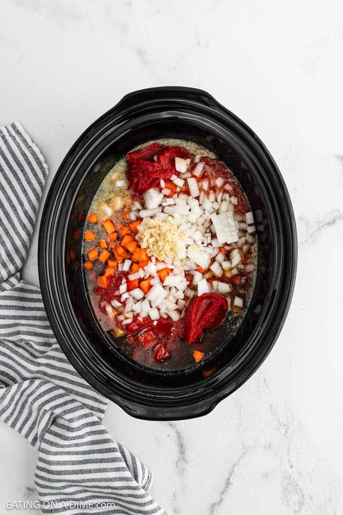 Overhead view of a black slow cooker containing the beginnings of a crock pot ground beef stew recipeโchopped onions, carrots, garlic, tomato paste, and diced tomatoesโresting beside a striped kitchen towel on a white surface.