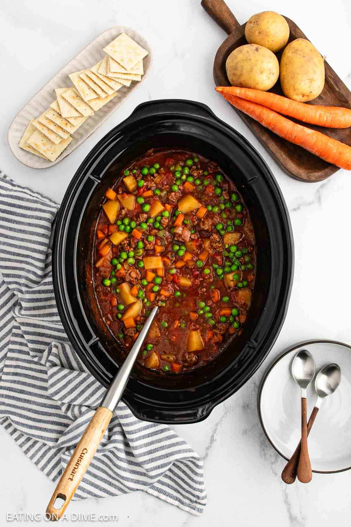 A crock pot ground beef stew recipe simmers in the slow cooker, filled with peas, carrots, and potatoes. Whole carrots, potatoes, crackers, and a plate with two spoons surround it on a striped towel atop a white surface.