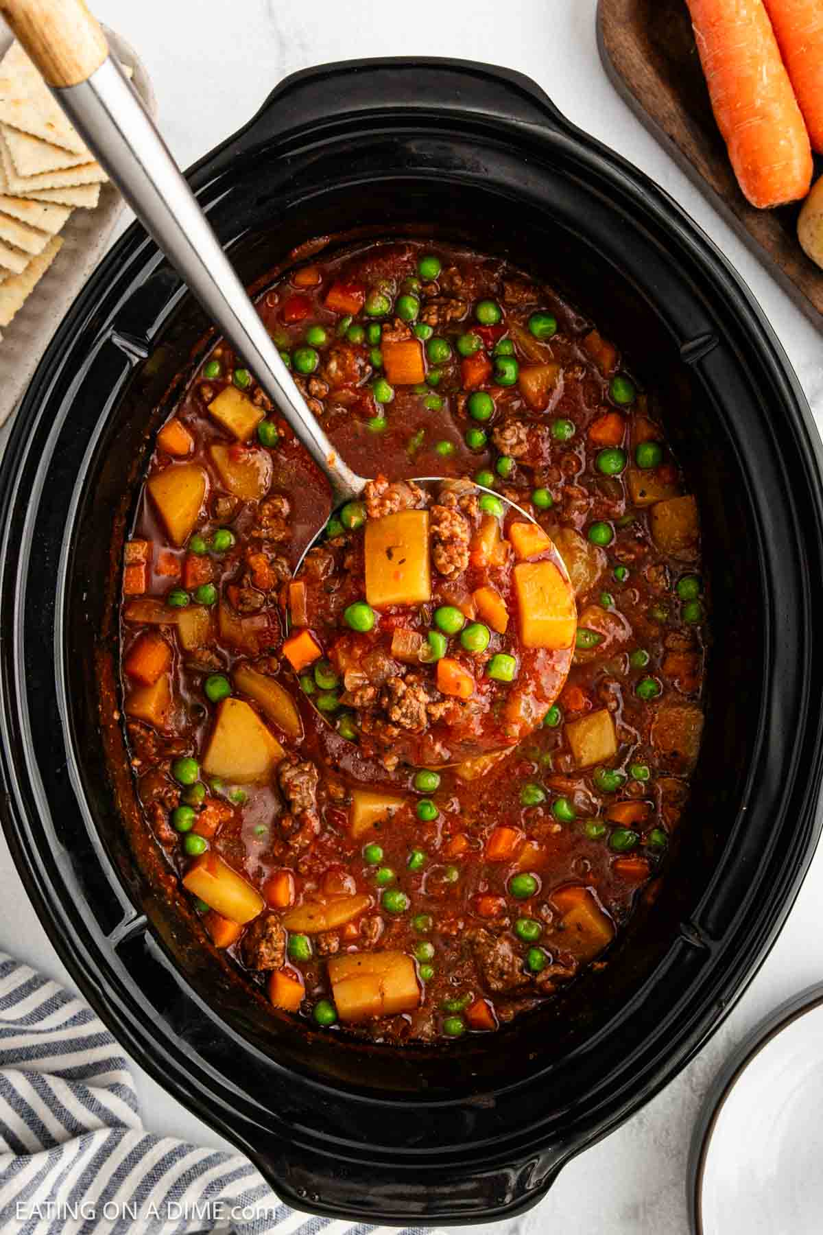 A ladle scoops hearty beef stew with potatoes, carrots, peas, and ground beef in a rich tomato broth from a black slow cookerโa delicious crock pot ground beef stew recipe. Crackers and fresh carrots sit on the side.