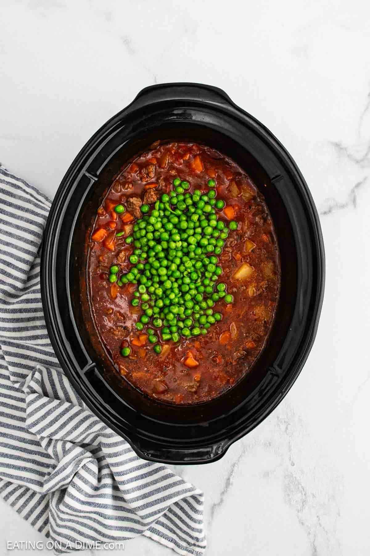 A crock pot ground beef stew recipe simmers in a slow cooker, topped with green peas and placed on a white marble surface next to a gray and white striped kitchen towel.