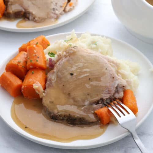A plate of food with sliced Instant Pot Pork Roast topped with gravy, mashed potatoes, and cooked carrots. A fork rests on the plate, spearing a carrot. Another plate of similar food is partially visible in the background.