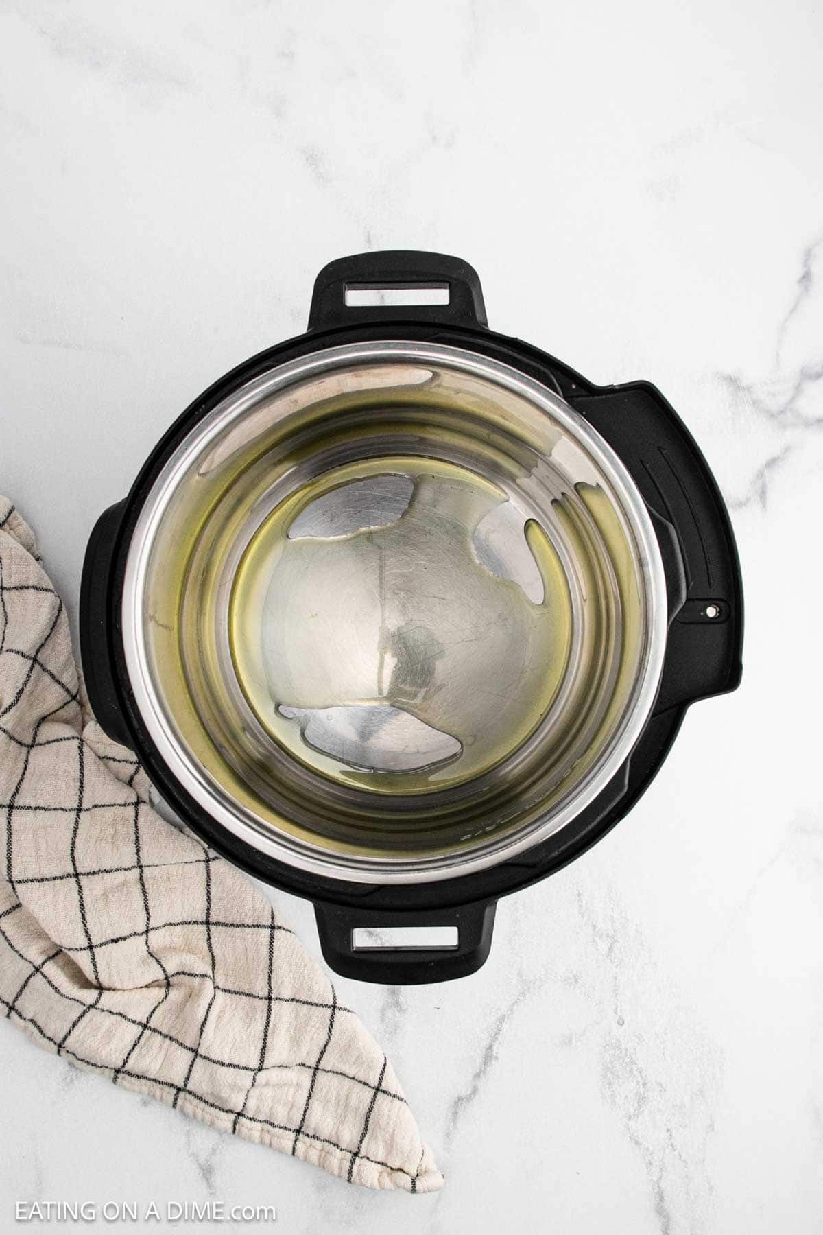 An overhead view of an empty Instant Pot with a small amount of oil inside, ready to begin cooking an Instant Pot Pork Roast, placed on a white marble surface next to a beige and black checkered kitchen towel.