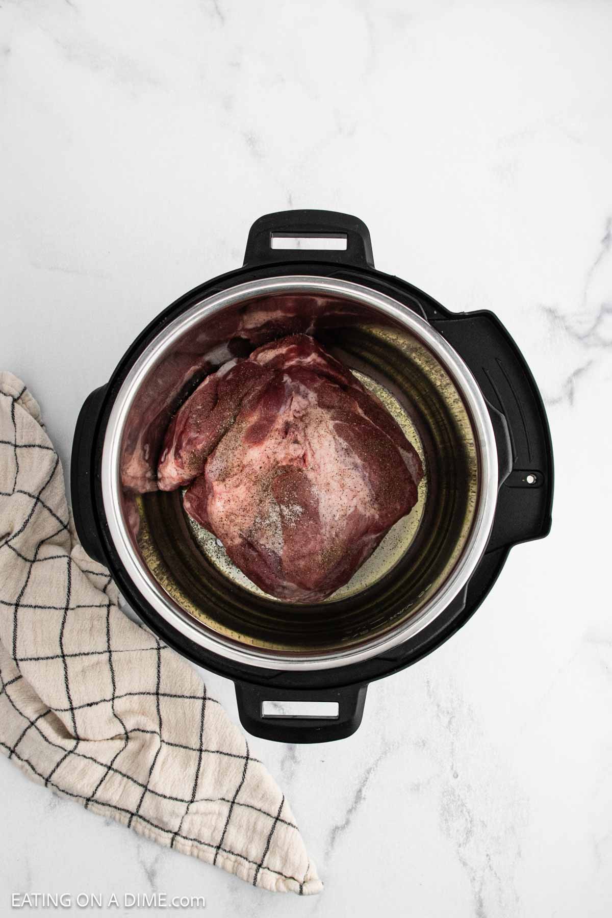 A raw pork shoulder, perfect for an Instant Pot Pork Roast, is seasoned with pepper and sits inside an open Instant Pot on a white marble surface, next to a beige and black checkered kitchen towel.