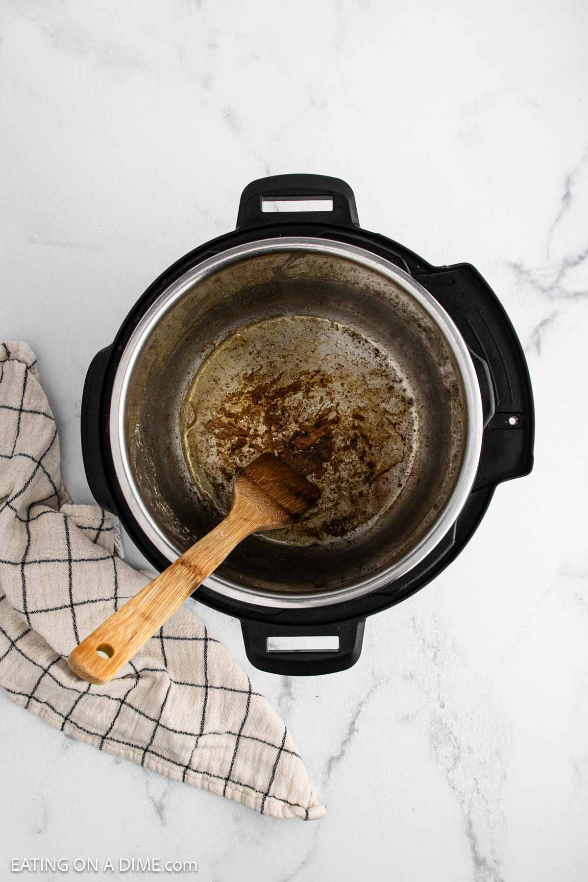An overhead view of an Instant Pot with browned bits from cooking Instant Pot Pork Roast and a wooden spatula inside. A white kitchen towel with black stripes rests beside the pot on a white marble surface.