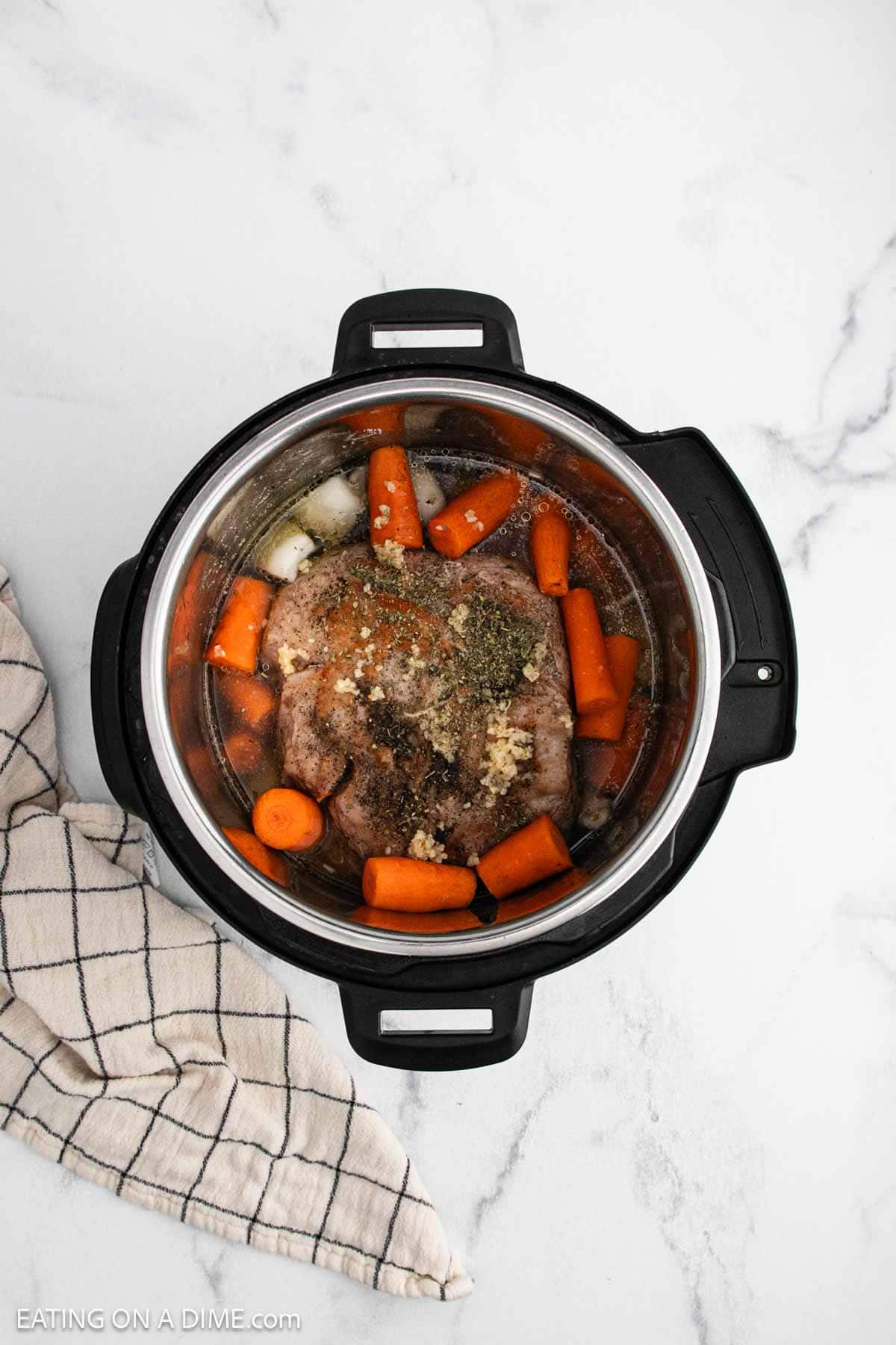 An overhead view of an Instant Pot Pork Roast with raw seasoned meat, whole carrots, onion pieces, and broth, sitting on a white marble surface next to a beige plaid kitchen towel.