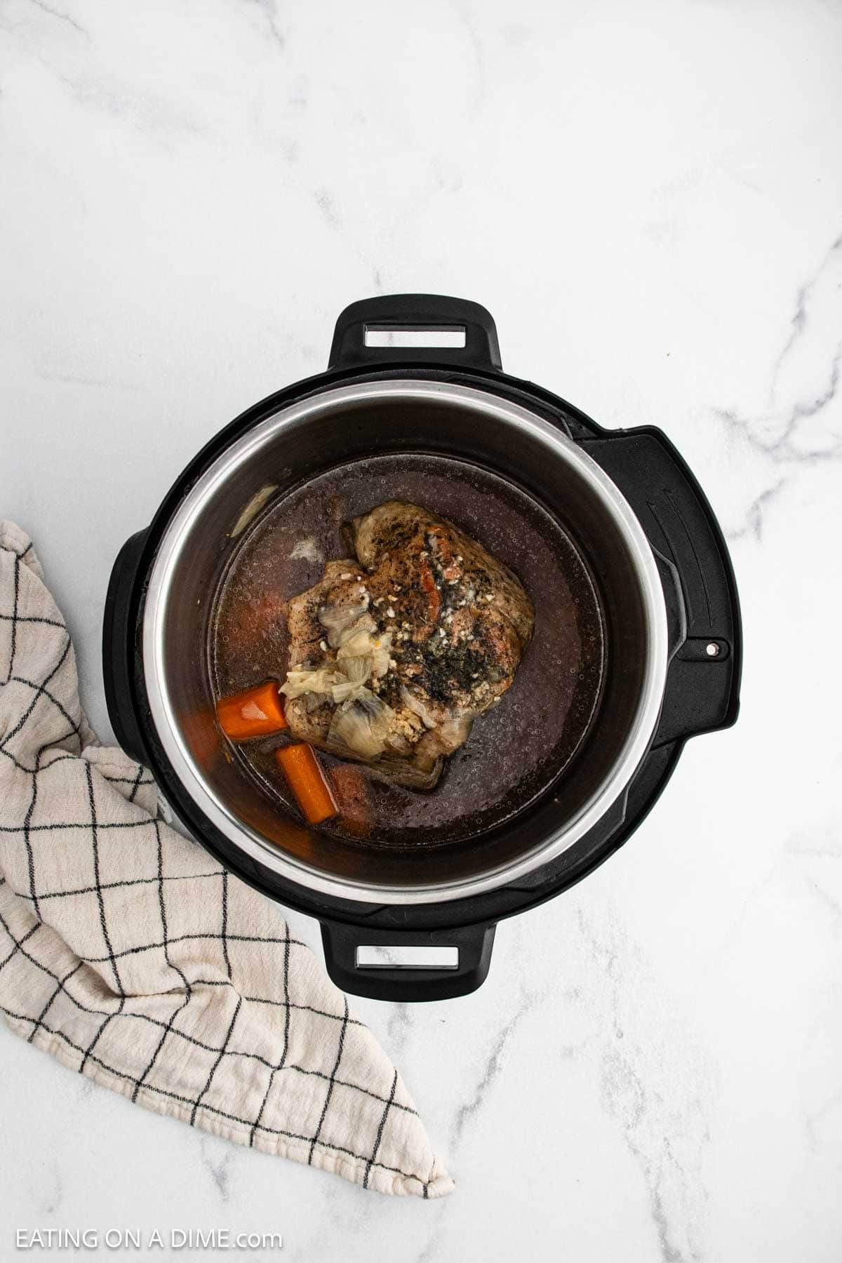 An overhead view of an Instant Pot Pork Roast with carrots and broth, cooked to perfection and placed on a white marble surface next to a beige and black checkered kitchen towel.