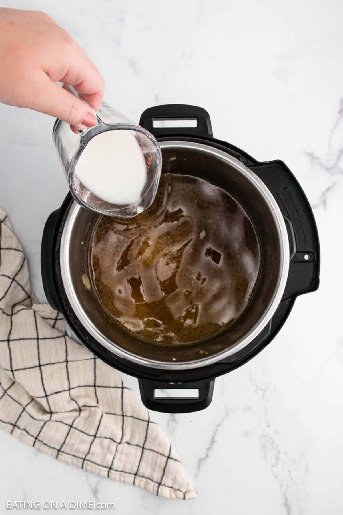 A hand pours milk from a glass measuring cup into a pot of broth for Instant Pot Pork Roast inside an electric pressure cooker on a marble countertop, next to a striped kitchen towel.