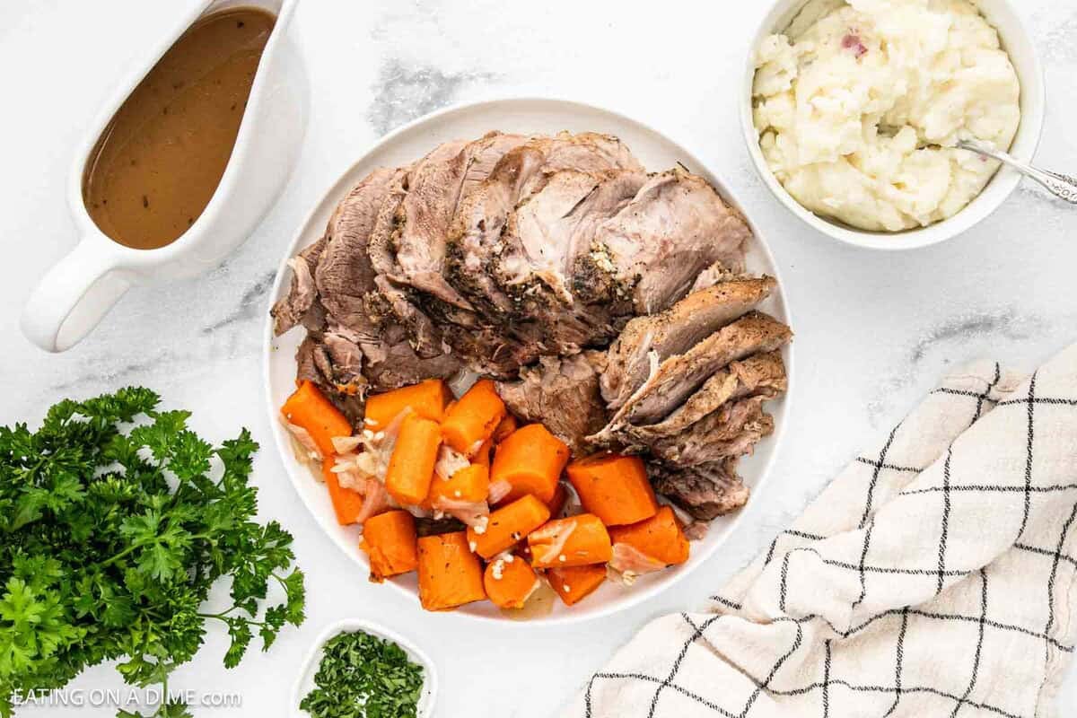 A plate with sliced Instant Pot Pork Roast, cooked carrots, and a side of mashed potatoes. A bowl of gravy, a bunch of parsley, and a checkered napkin are nearby on a white surface.