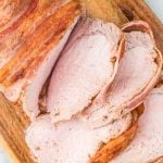 Close-up of several slices of juicy Bacon Wrapped Pork Loin on a wooden cutting board, showing the tender, light pink meat and golden-brown bacon crust.