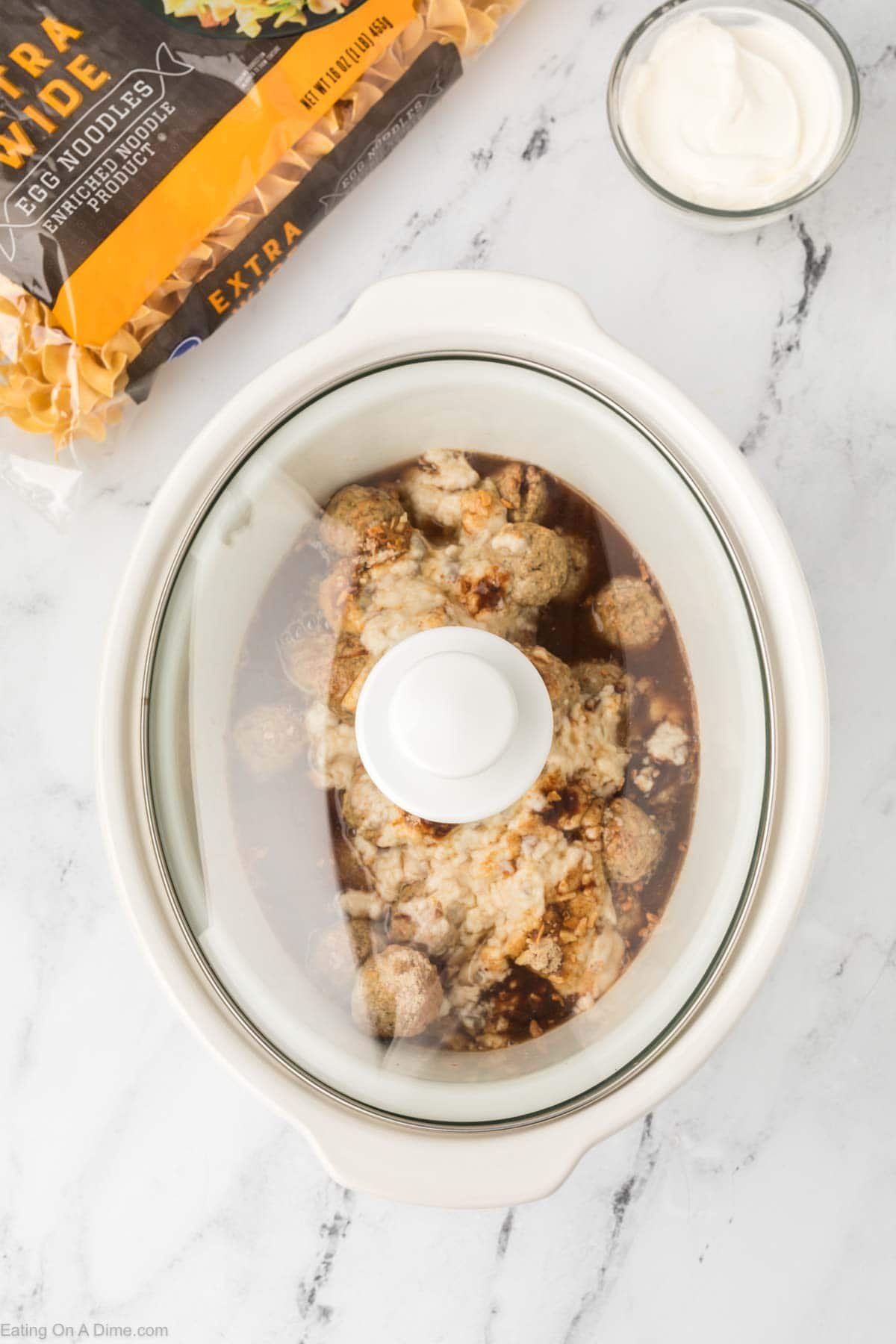 A Crock Pot Swedish Meatballs dish in a slow cooker sits on a marble countertop, smothered in brown sauce and creamy topping. Nearby are a bag of extra wide egg noodles and a bowl of sour cream for serving.