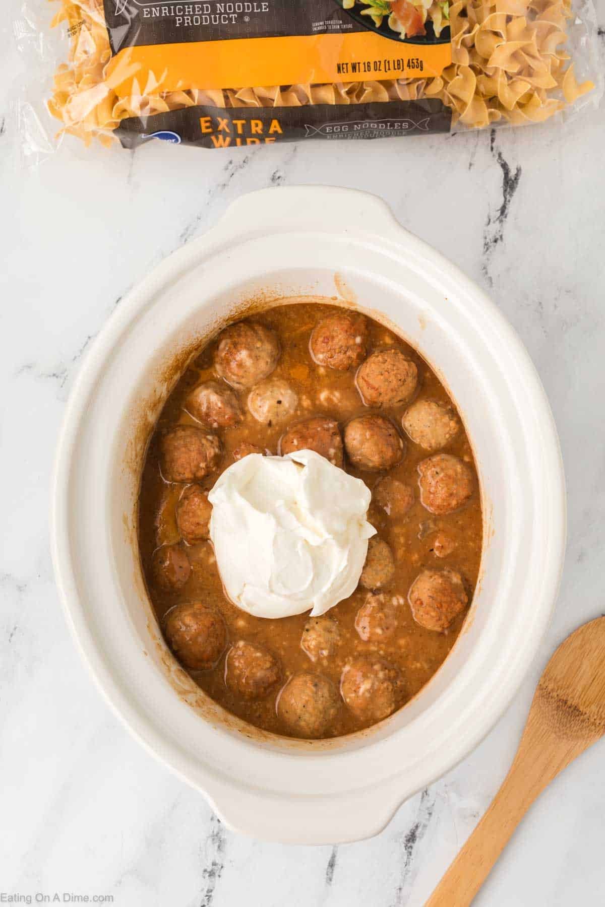 A white Crock Pot filled with Swedish Meatballs in brown sauce, topped with a dollop of sour cream. A wooden spoon and a package of uncooked egg noodles rest on the marble countertop.
