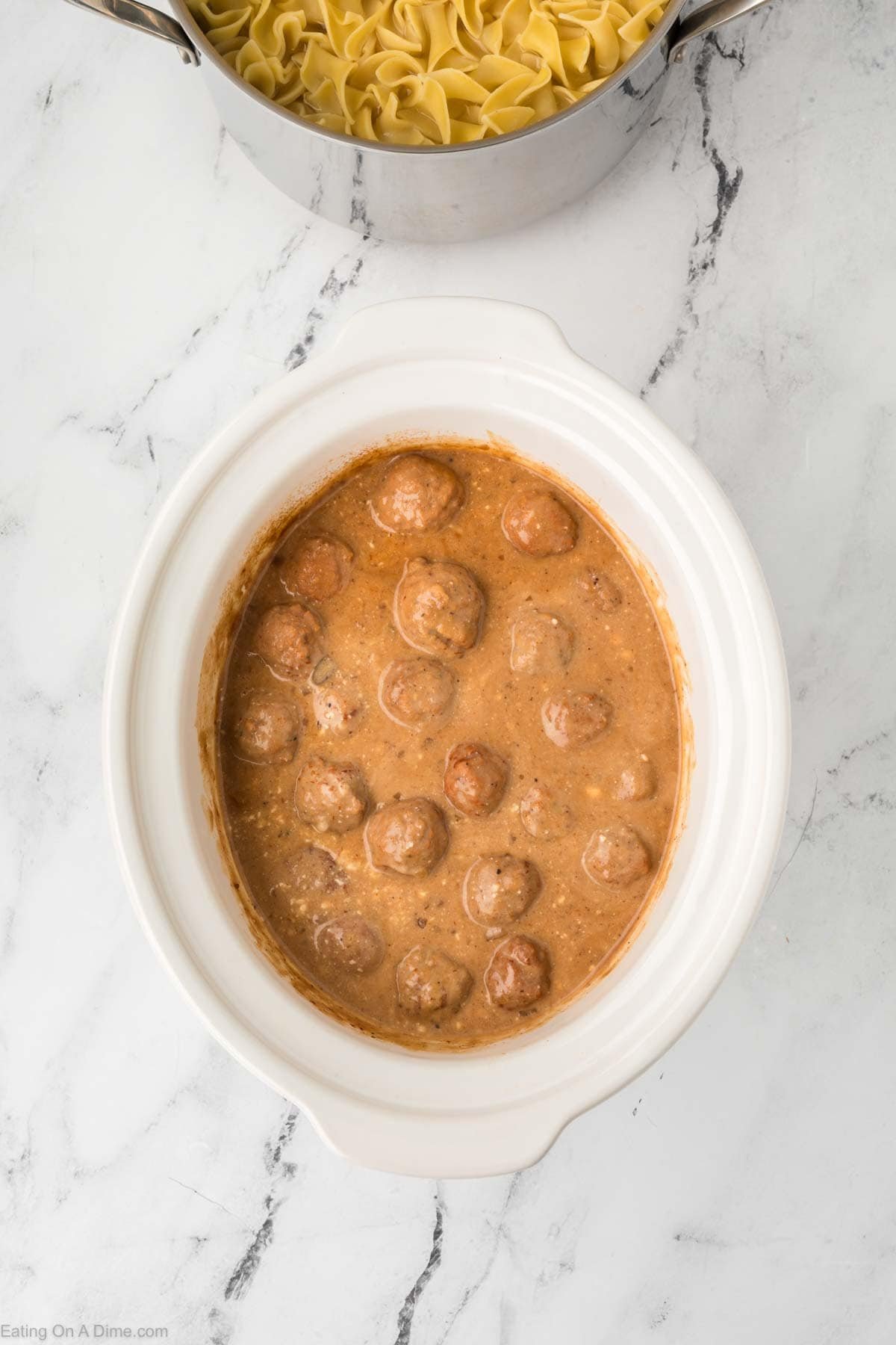 A white oval slow cooker filled with Crock Pot Swedish Meatballs in creamy brown sauce sits on a marble countertop. Above it, a pot of uncooked egg noodles is partially visible.