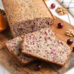 A loaf of cranberry walnut bread on a wooden board, with two slices cut in front. Cranberry pieces and walnuts are visible in the bread, reminiscent of a classic cranberry orange bread recipe, with whole cranberries and nuts scattered nearby.