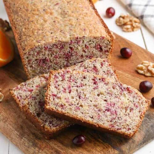 A loaf of cranberry walnut bread on a wooden board, with two slices cut in front. Cranberry pieces and walnuts are visible in the bread, reminiscent of a classic cranberry orange bread recipe, with whole cranberries and nuts scattered nearby.