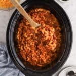 A wooden spoon lifts a serving of Crock Pot Chili Mac and Cheese Recipe—cheesy macaroni and ground beef in tomato sauce—from a black slow cooker. A bowl of shredded cheese and a striped towel are nearby.