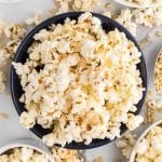 A black bowl filled with freshly popped Kettle Corn Popcorn, surrounded by scattered popcorn kernels and pieces on a light-colored surface.