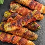 Several bacon wrapped pickles and jalapeño poppers are arranged on a dark slate surface, showcasing a tempting mix of melted cheese and perfectly charred bacon.