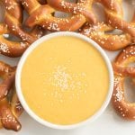A white bowl of creamy Beer Cheese Dip topped with sprinkled salt sits surrounded by soft pretzels with coarse salt on a white surface.