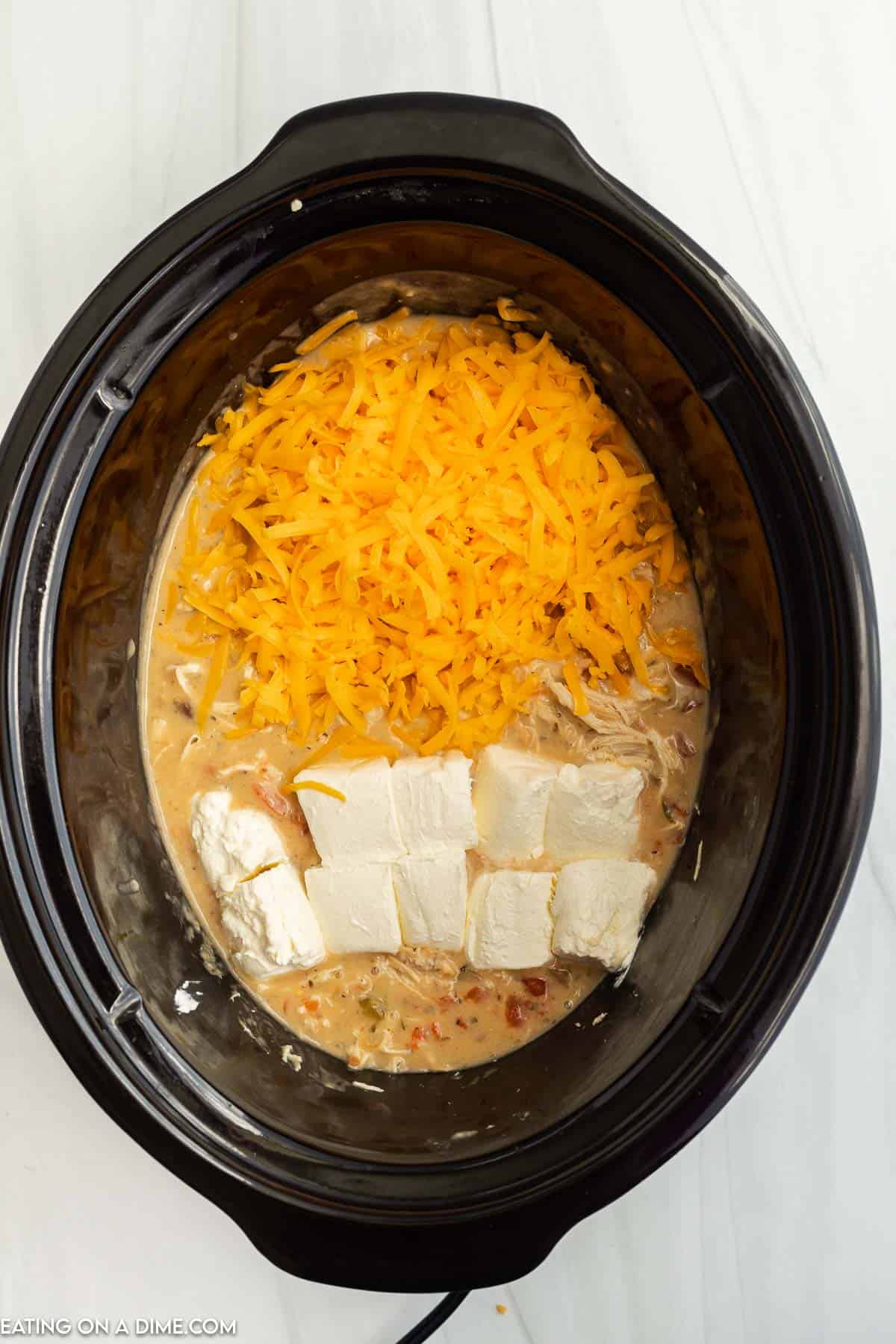 Topping the cooked chicken mixture with shredded cheese and cubed cream cheese in the slow cooker