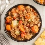 A bowl of hearty Crock Pot Poor Man's Stew with ground beef, diced potatoes, carrots, onions, and tomatoes, garnished with herbs. A slice of cornbread and utensils are visible nearby on a light-colored table.