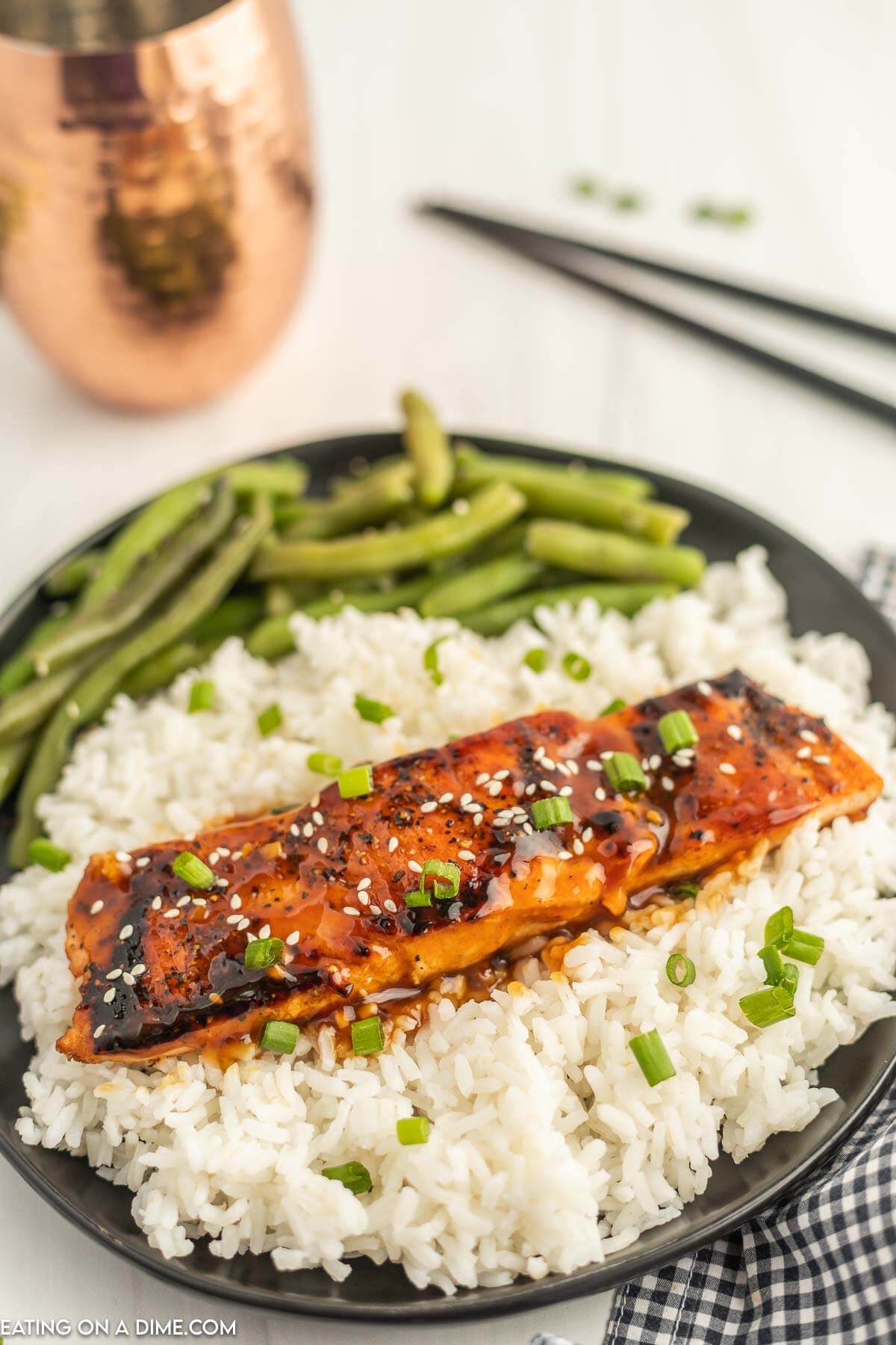 Teriyaki Salmon on a black plate topped with sesame seeds and diced green onions. Topped on white rice and a side of green beans