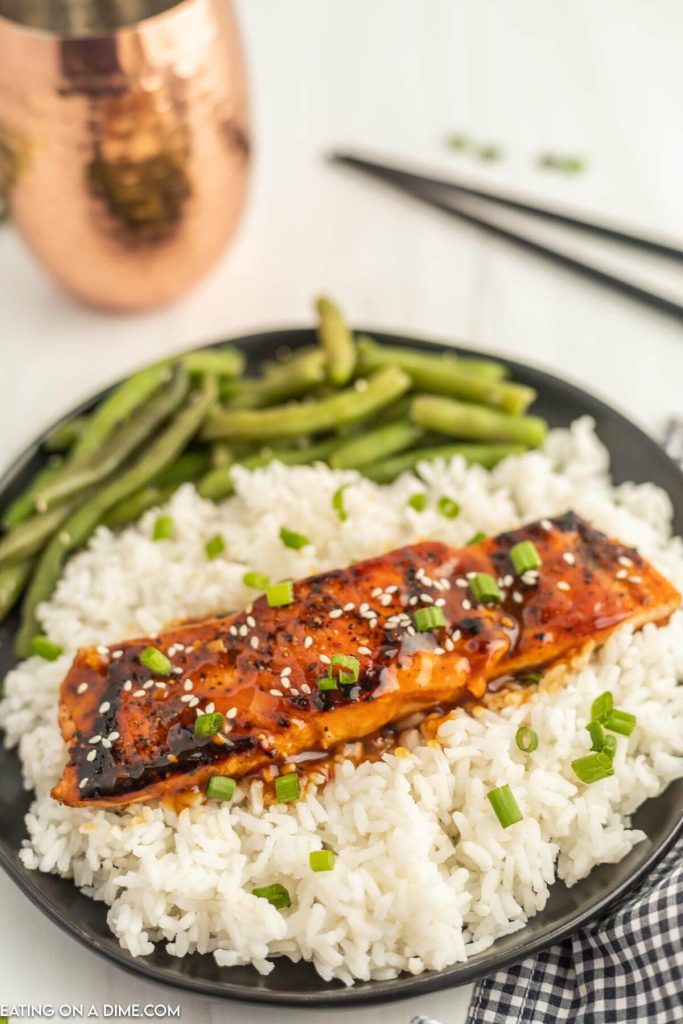 Teriyaki Salmon on a black plate topped with sesame seeds and diced green onions. Topped on white rice and a side of green beans