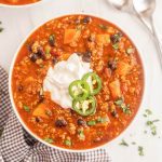 A bowl of turkey sweet potato chili topped with sour cream and sliced jalapeños. The chili features black beans, diced tomatoes, and sweet potato chunks, garnished with chopped cilantro. Two spoons and a checkered napkin are nearby.