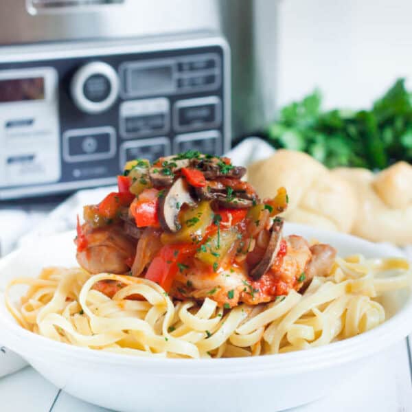 A dish of pasta topped with a blend of slow cooker chicken cacciatore, mushrooms, and peppers in a savory sauce, garnished with herbs. Fresh parsley is blurred in the background. Bread rolls are visible on the side.