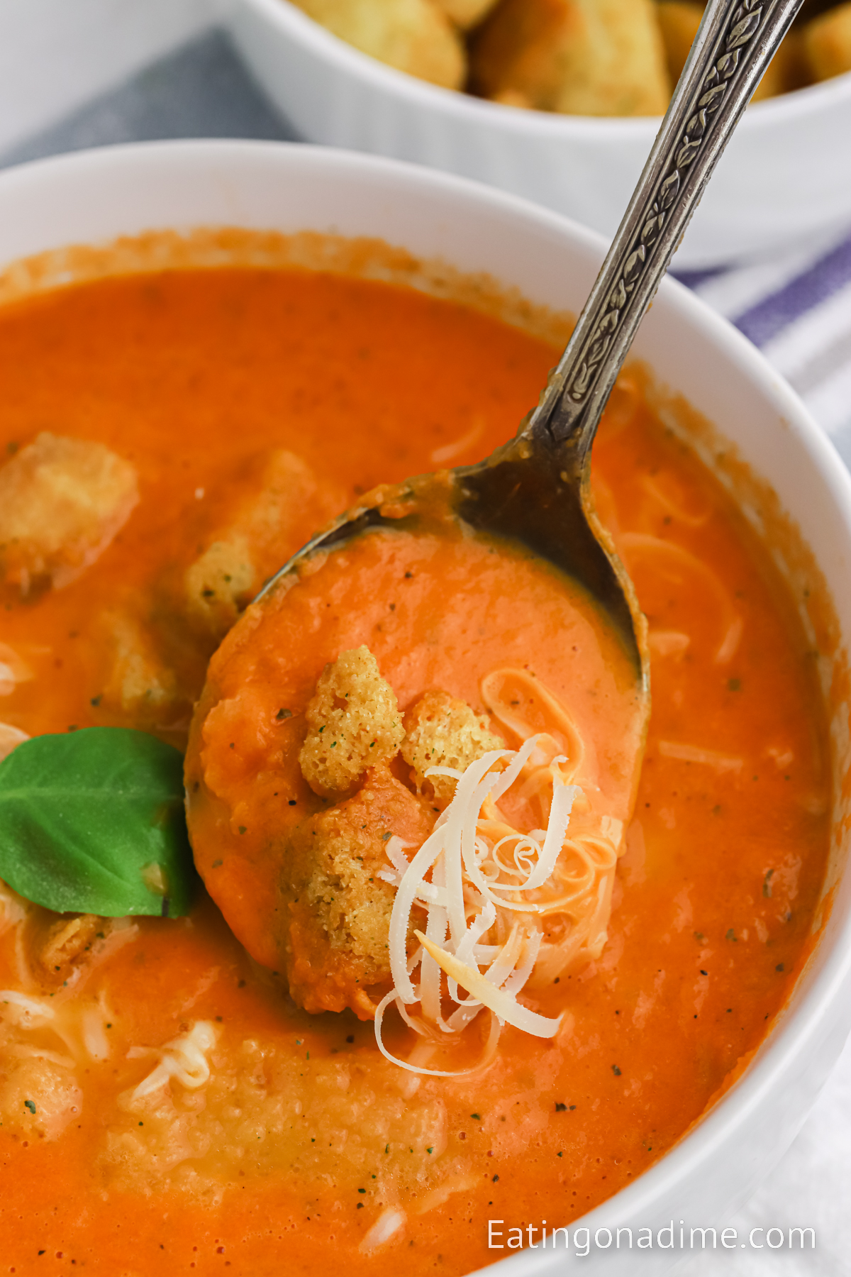 A bowl of tomato soup topped with croutons, fresh basil and cheese with a serving on a spoon