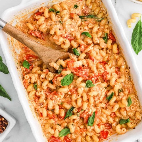 A white baking dish filled with baked pasta, cherry tomatoes, crumbled feta cheese, and fresh basil leaves, with a wooden spoon stirring the trending Tiktok Feta Pasta mixture.