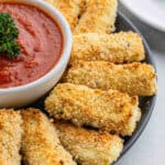 A plate of crispy homemade mozzarella cheese sticks arranged around a bowl of marinara sauce, garnished with a sprig of parsley.