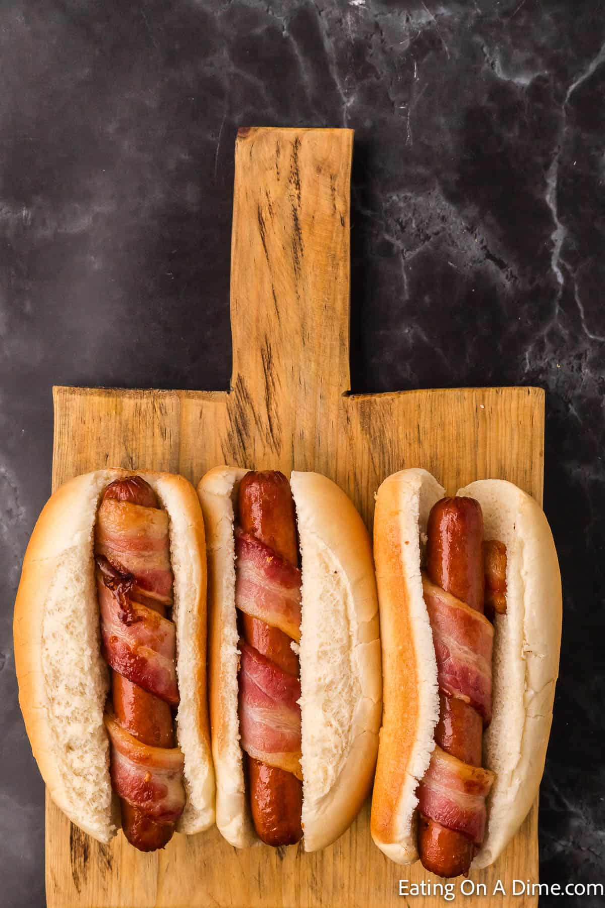 Bacon wrapped hot dogs in hot dog buns on a cutting board