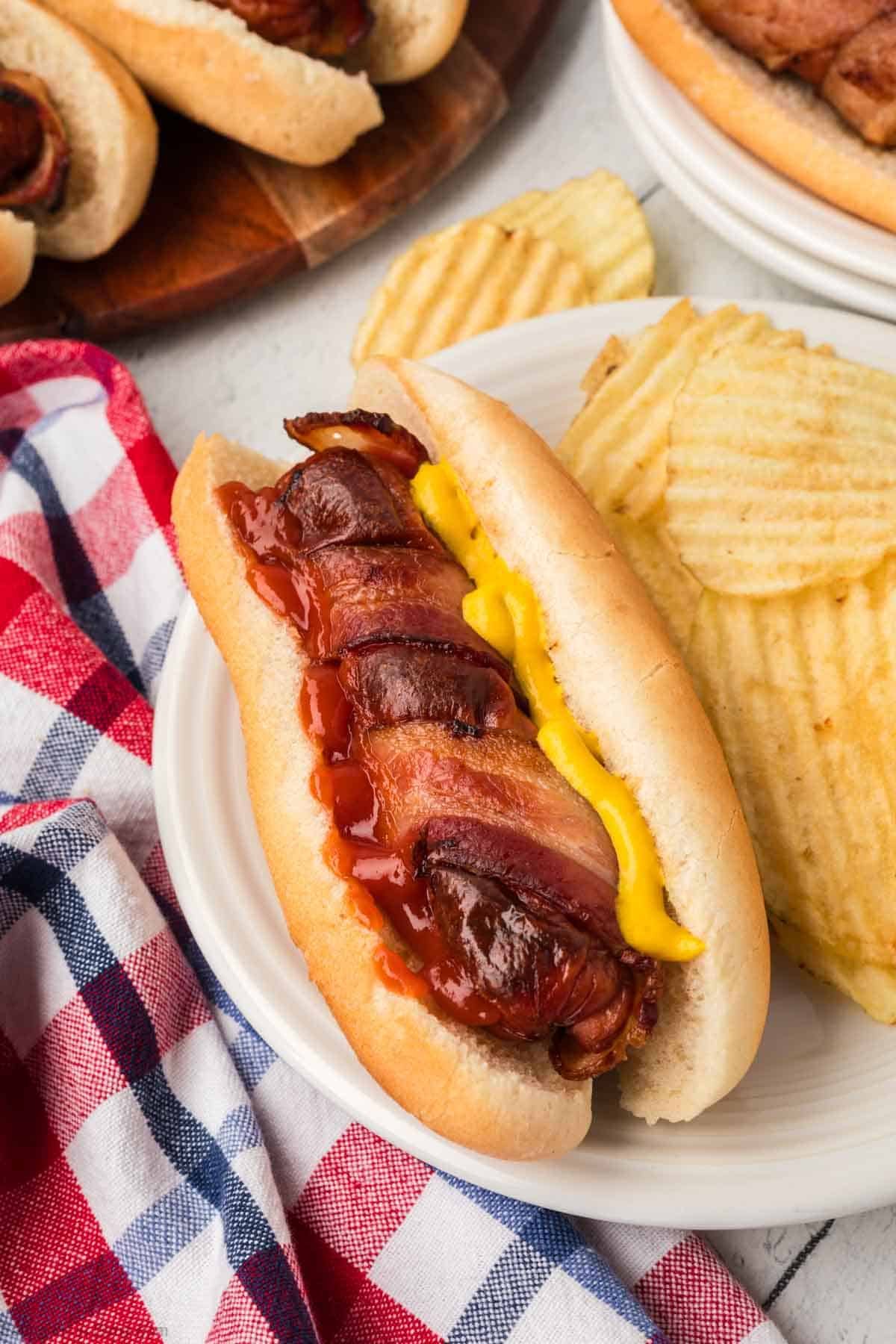 Bacon Wrapped Hot Dogs topped with mustard and ketchup with a side of potato chips