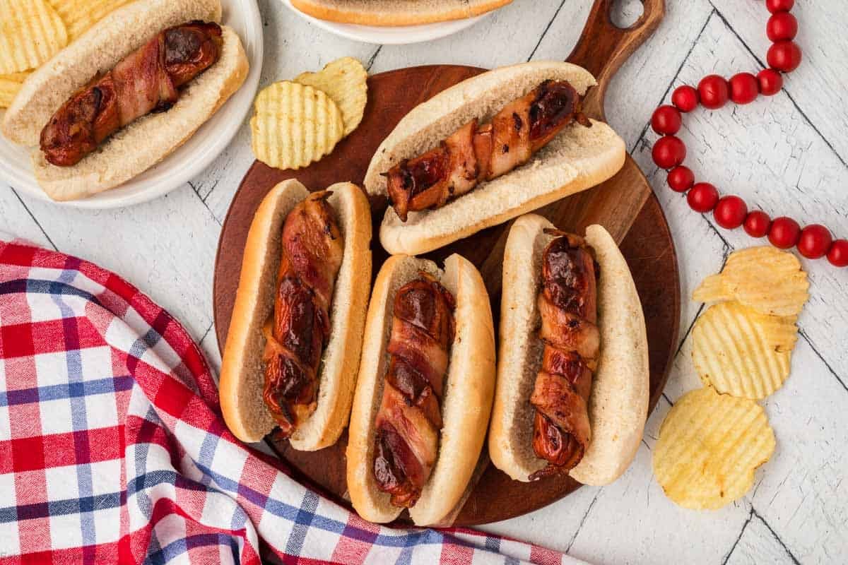 Bacon wrapped hot dogs on a wooden platter with potato chips on the side