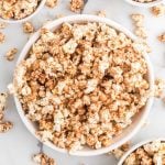 A white bowl filled with cinnamon popcorn, caramel-coated to perfection, sits on a marble surface with more popcorn scattered around and partially visible bowls in the corners.