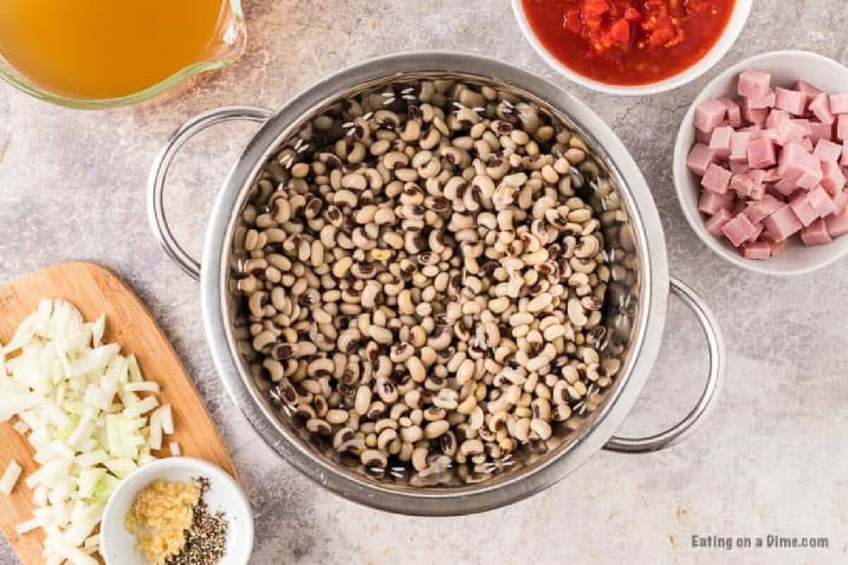 A colander filled with black-eyed peas sits on a counter surrounded by bowls of chopped ham, diced tomatoes, onions, garlic with spices, and broth—everything you need for savory Crock Pot Black Eyed Peas.