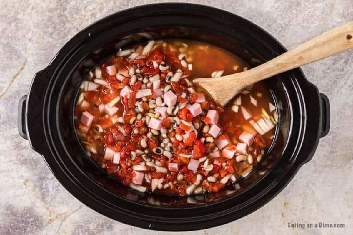 A Crock Pot Black Eyed Peas dish simmers with diced ham, chopped onions, tomatoes, and broth, all blended together in a slow cooker with a wooden spoon resting inside.