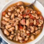 A white bowl filled with Crock Pot Black Eyed Peas, cooked with diced tomatoes, onions, and chunks of meat in a savory broth, with a spoon resting in the bowl.