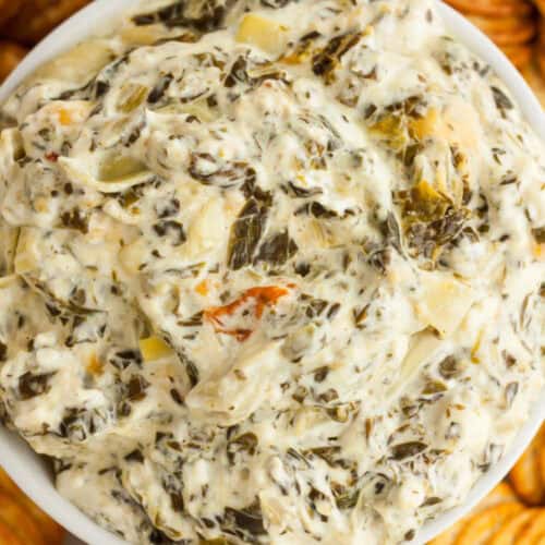 A close-up of a bowl filled with creamy Crockpot Spinach Artichoke Dip, showing chunks of vegetables mixed into the smooth, cheesy mixture. Crackers are visible around the bowl’s edge.