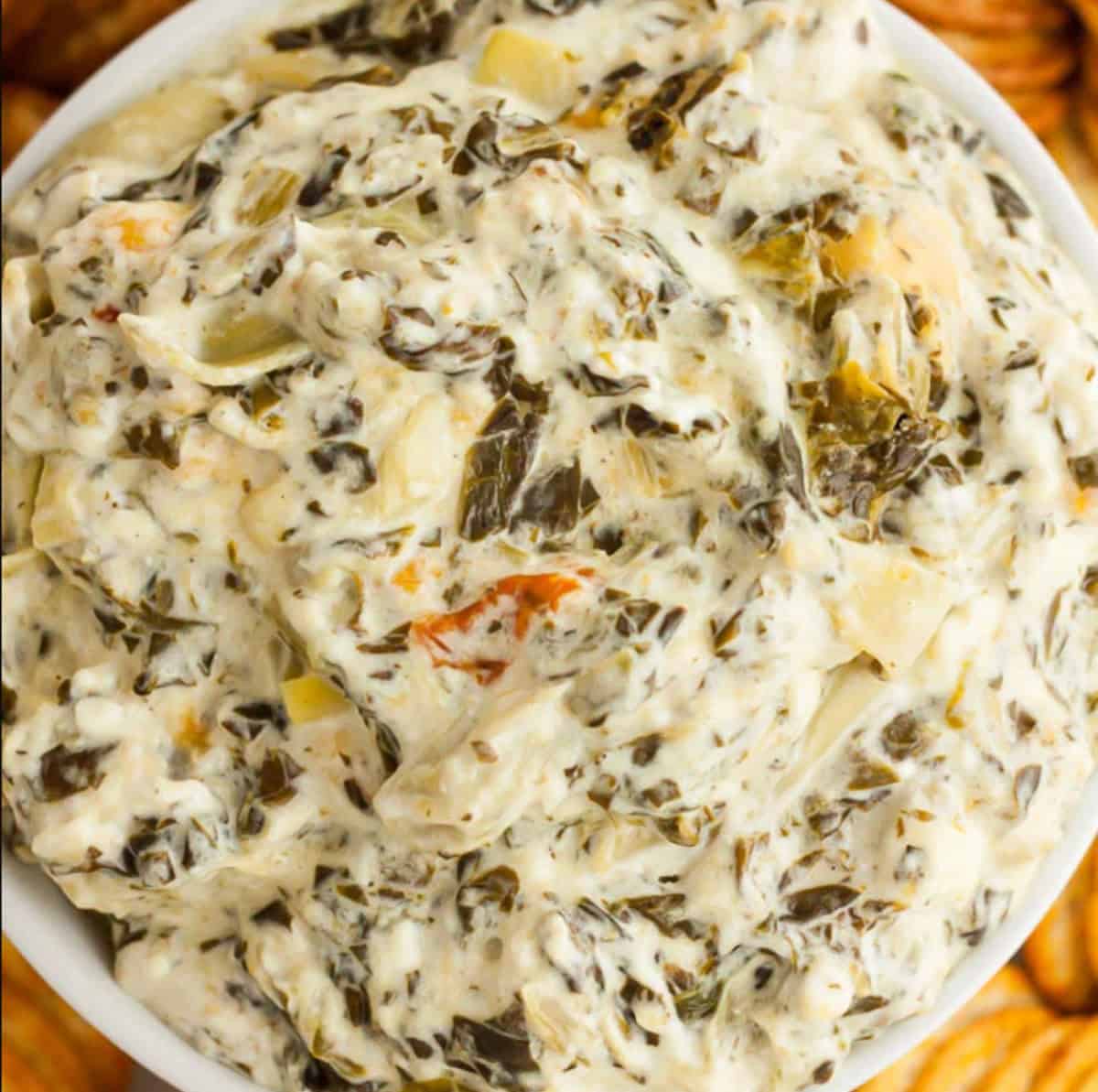 A close-up of a bowl filled with creamy Crockpot Spinach Artichoke Dip, showing chunks of vegetables mixed into the smooth, cheesy mixture. Crackers are visible around the bowl’s edge.