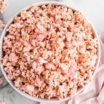 A white bowl filled with Pink Popcorn sits on a marble surface, with a pink and white striped cloth nearby and a few pieces of Pink Popcorn scattered around.