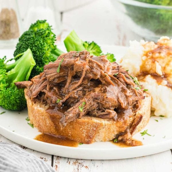 Shredded Slow Cooker Roast Beef Sandwiches with savory gravy served on thick bread, paired with steamed broccoli and creamy mashed potatoes smothered in gravy on a white plate.