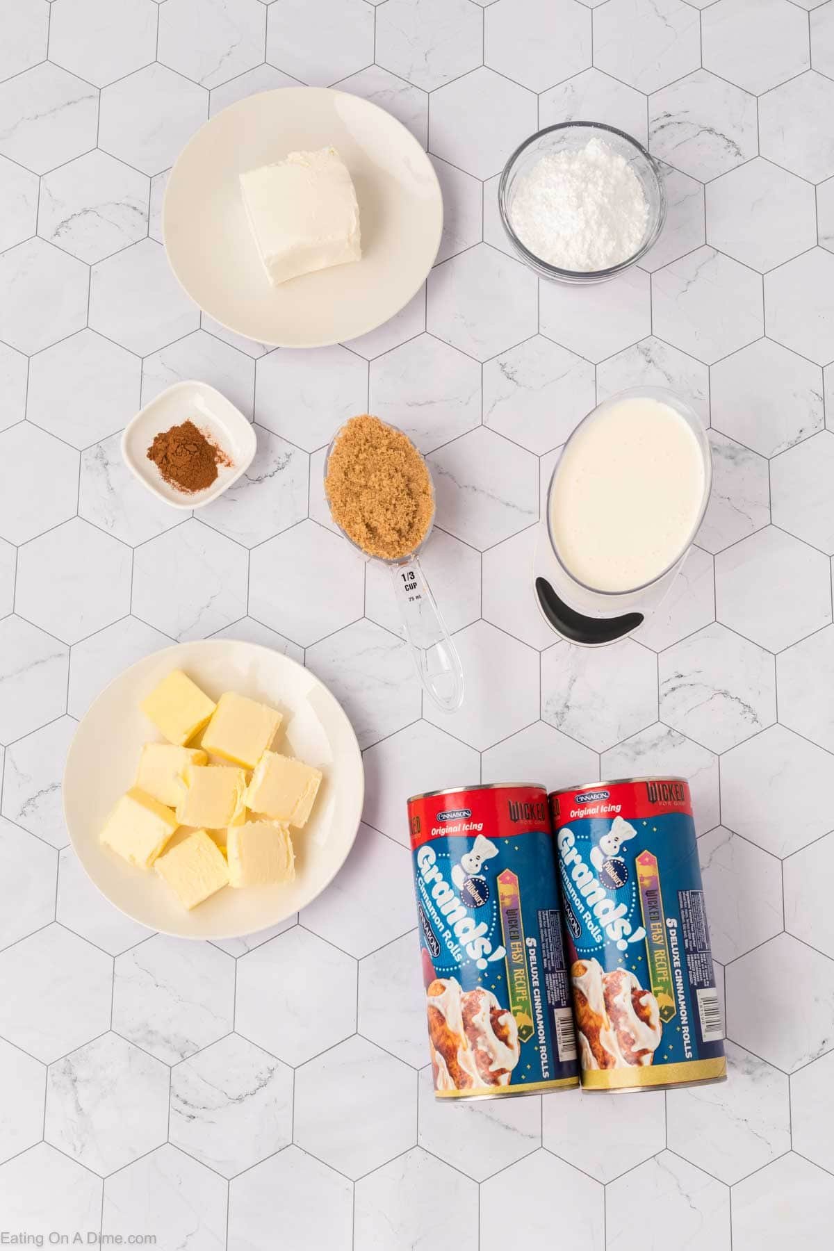 Ingredients for a TikTok Cinnamon Rolls Recipe are arranged on a white hexagon-tiled surface, including cream cheese, powdered sugar, brown sugar, butter, cinnamon, heavy cream, and two cans of refrigerated cinnamon rolls.
